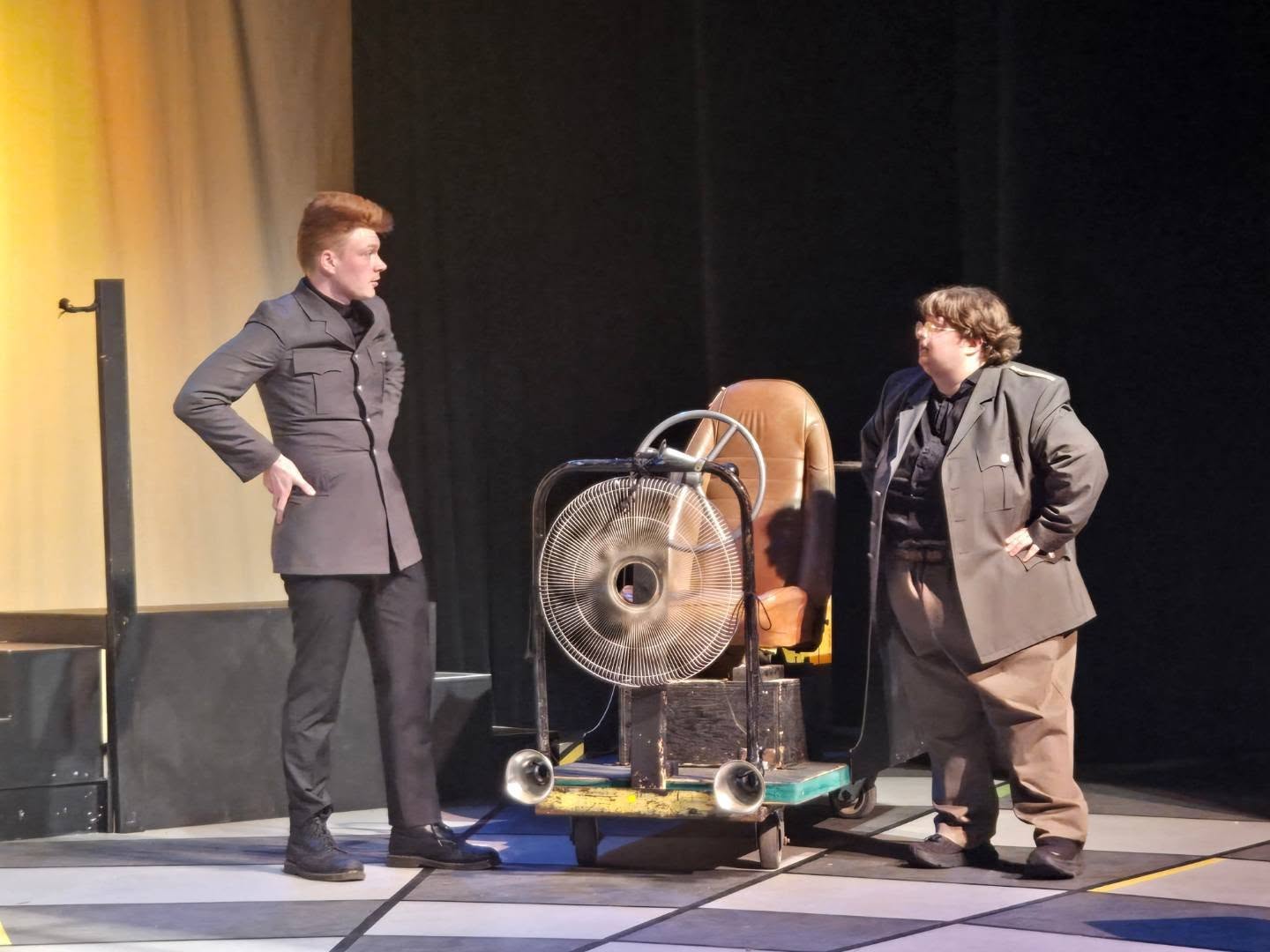 Two performers beside a wheeled prop assembled with a chair, fan, and steering wheel