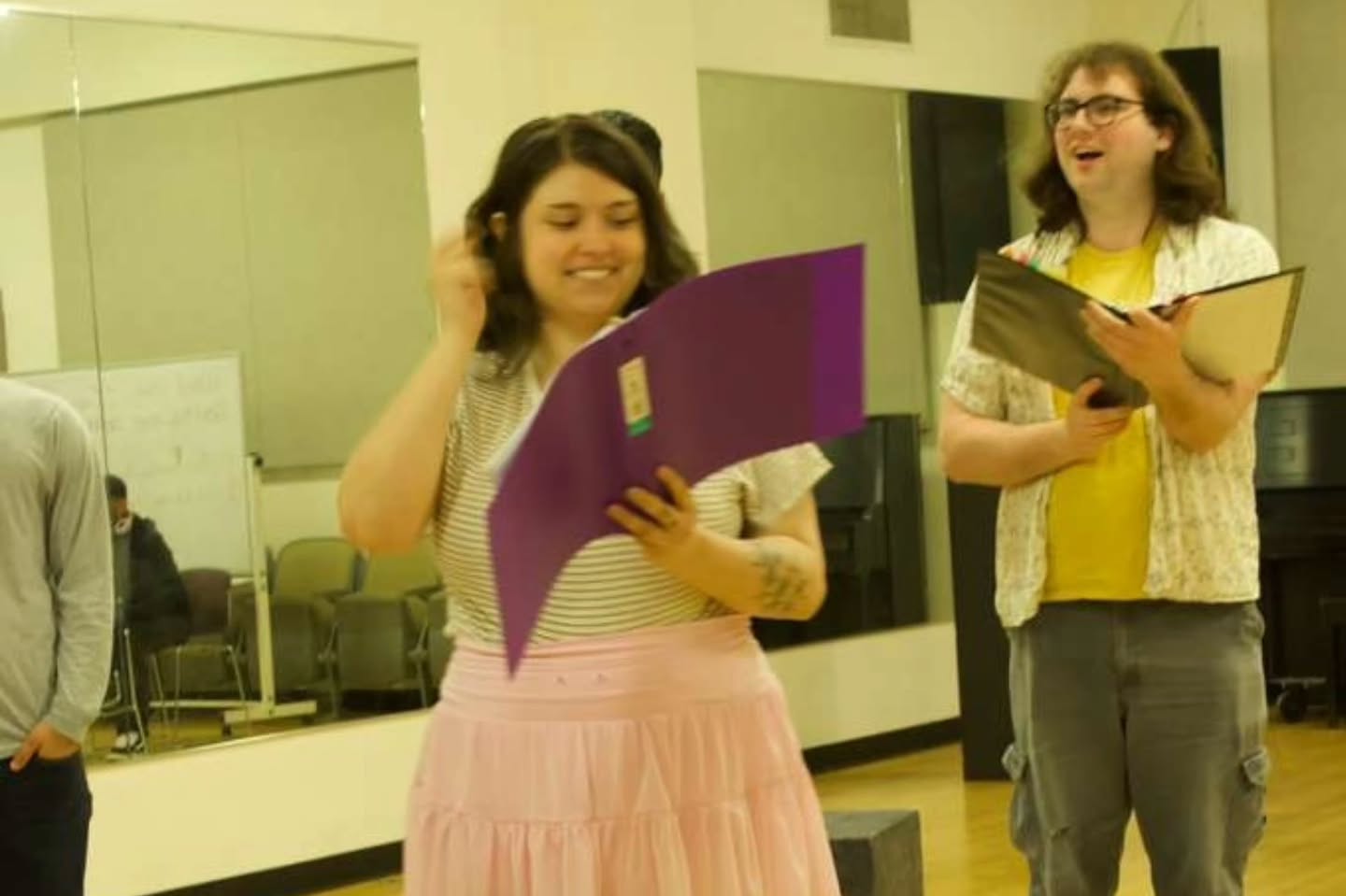 Two performers smiling while reading from script folders in the rehearsal room
