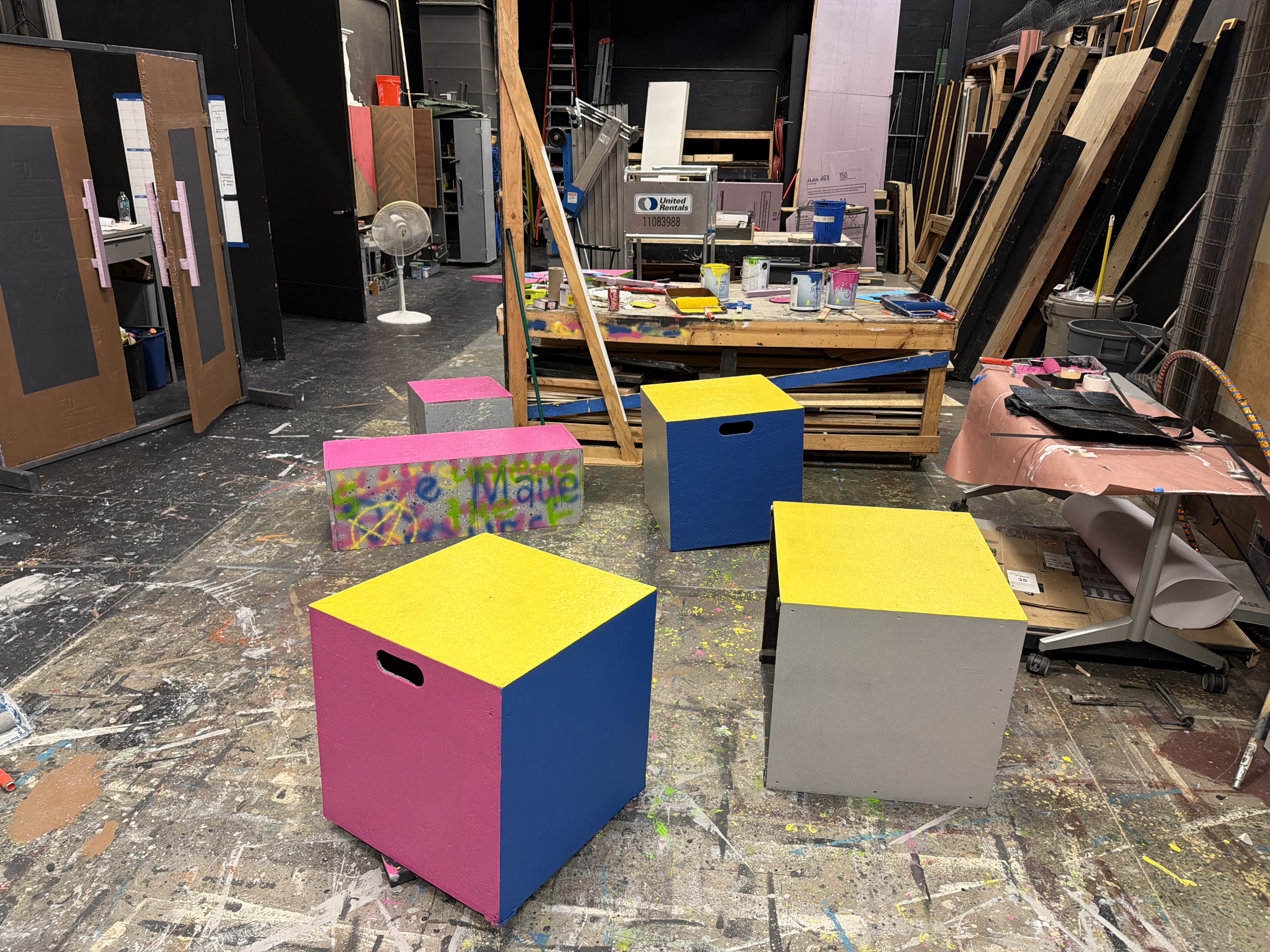 Brightly painted pink, yellow, and blue set pieces beside a paint-covered workbench in the scene shop
